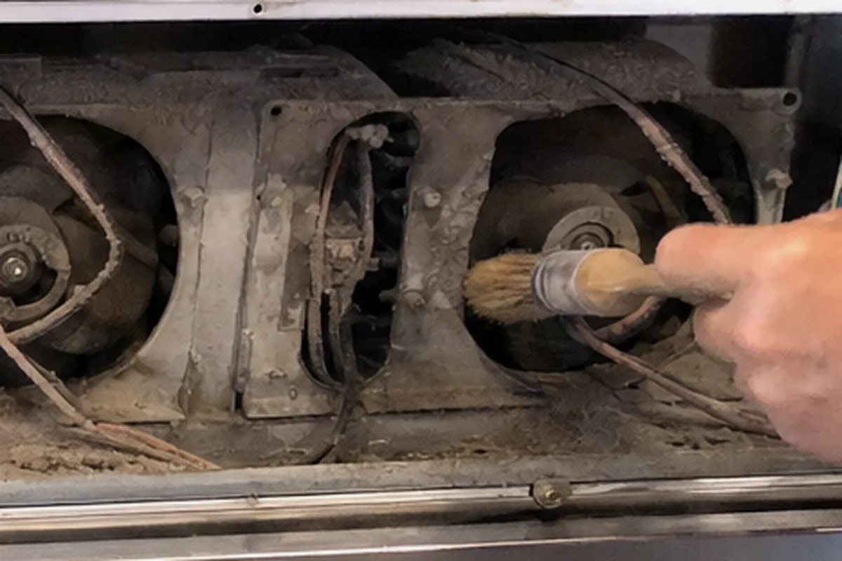 A soft brush being drawn across a dust-clogged fan and coil bank inside an opened appliance compartment